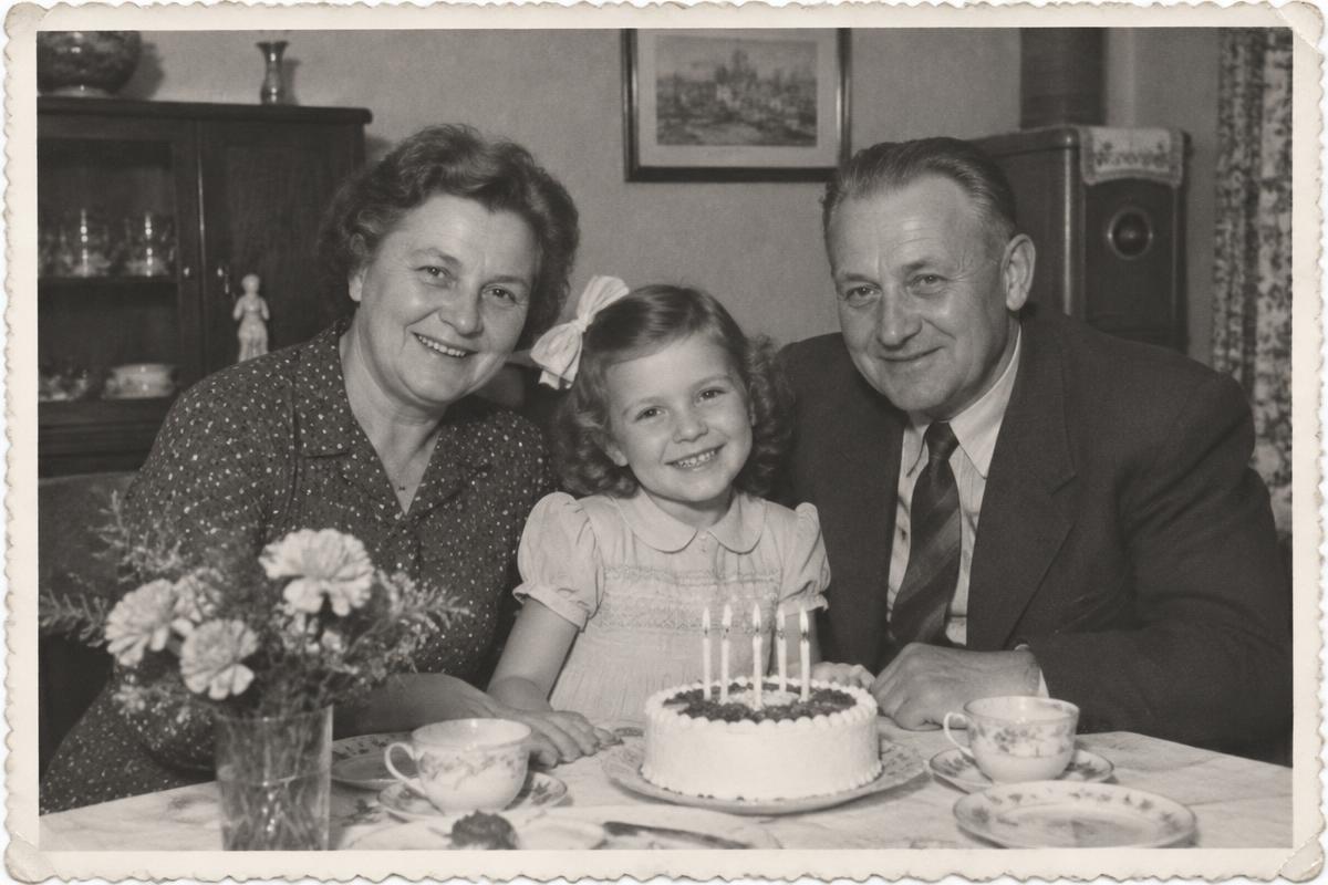 Ilustrační černobílá fotografie ve stylu 50. let: prarodiče s vnučkou u narozeninového dortu.